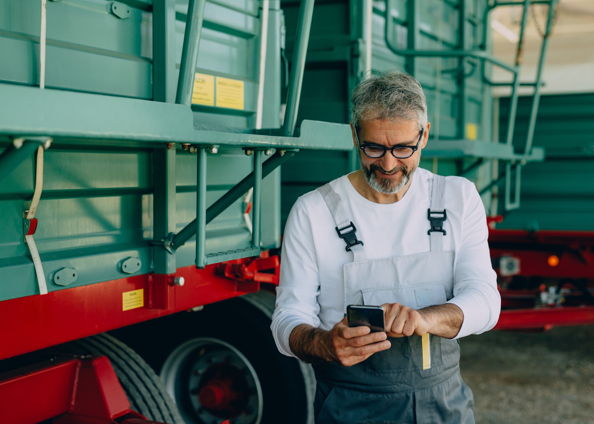 A farmers standing beside a green agricultural trailer, smiling as he checks something on his smartphone.